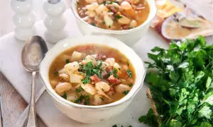 Dwie miseczki sycącej zupy na rosole z fasolą, ziemniakami i kiełbasą, posypane natką pietruszki.