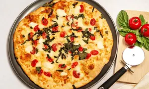 Pyszna pizza z serka wiejskiego, pomidorkami koktajlowymi i bazylią. Obok leży krajalnica do pizzy i świeże pomidory.