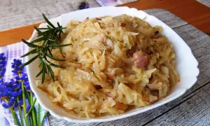 Młoda kapusta zasmażana z boczkiem, podana w białej misce, ozdobiona gałązką rozmarynu.