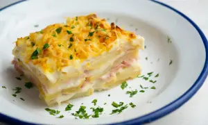 Kawałek pysznej, zapiekanej lasagne ziemniaczanej z szynką i serem, posypany natką pietruszki.