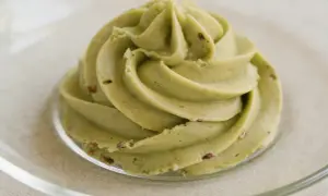 Zielona, kremowa masa pistacjowa z kawałkami pistacji, uformowana w kształt róży na szklanym talerzyku.