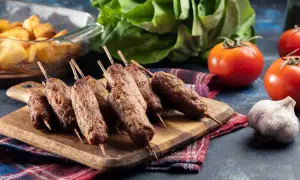 Grillowane ćevapi na drewnianej desce, obok pomidorów, czosnku i pieczonych ziemniaków.
