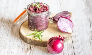 Słoik z konfiturą z czerwonej cebuli, obok cała cebula i połówka. Gałązka rozmarynu dodaje aromatu.