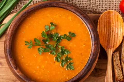 Aksamitna zupa pomidorowa w drewnianej misce, udekorowana natką pietruszki. Obok drewniana łyżka i kromki chleba. Idealna do przyprawienia na wiele sposobów.