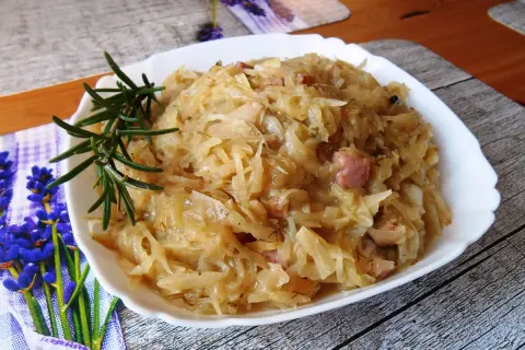 Młoda kapusta zasmażana z boczkiem, podana w białej misce, ozdobiona gałązką rozmarynu.