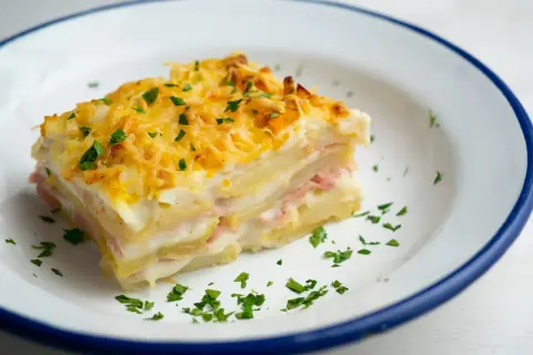 Kawałek pysznej, zapiekanej lasagne ziemniaczanej z szynką i serem, posypany natką pietruszki.