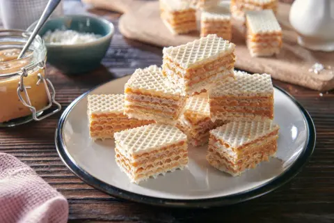 Słodkie wafle przełożone kremową masą do wafli, ułożone na talerzu. Obok słoik z masą i miseczka z wiórkami.