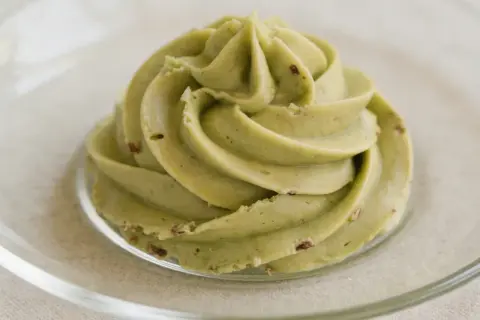 Zielona, kremowa masa pistacjowa z kawałkami pistacji, uformowana w kształt róży na szklanym talerzyku.