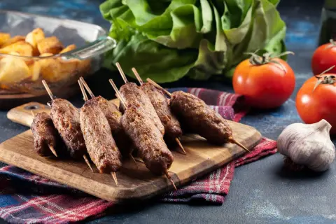 Grillowane ćevapi na drewnianej desce, obok pomidorów, czosnku i pieczonych ziemniaków.