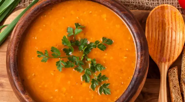 Aksamitna zupa pomidorowa w drewnianej misce, udekorowana natką pietruszki. Obok drewniana łyżka i kromki chleba. Idealna do przyprawienia na wiele sposobów.