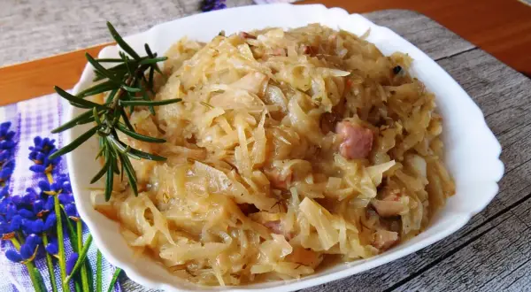 Młoda kapusta zasmażana z boczkiem, podana w białej misce, ozdobiona gałązką rozmarynu.
