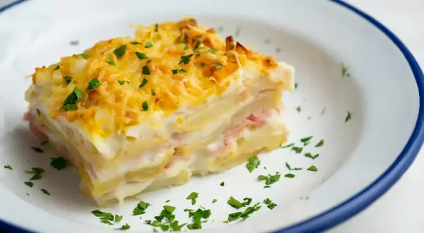 Kawałek pysznej, zapiekanej lasagne ziemniaczanej z szynką i serem, posypany natką pietruszki.