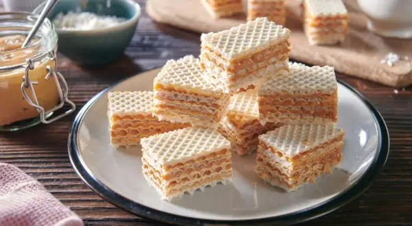 Słodkie wafle przełożone kremową masą do wafli, ułożone na talerzu. Obok słoik z masą i miseczka z wiórkami.