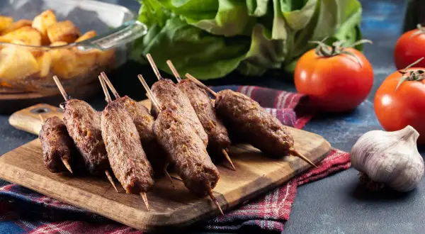 Grillowane ćevapi na drewnianej desce, obok pomidorów, czosnku i pieczonych ziemniaków.