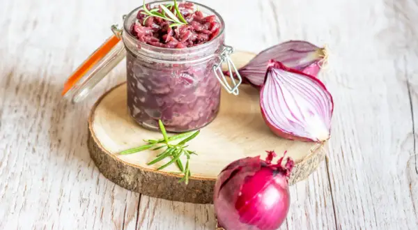 Słoik z konfiturą z czerwonej cebuli, obok cała cebula i połówka. Gałązka rozmarynu dodaje aromatu.