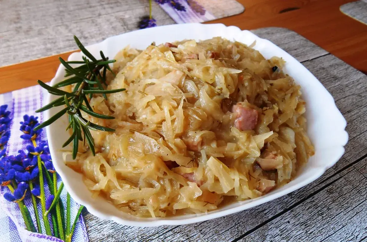 Młoda kapusta zasmażana z boczkiem, podana w białej misce, ozdobiona gałązką rozmarynu.