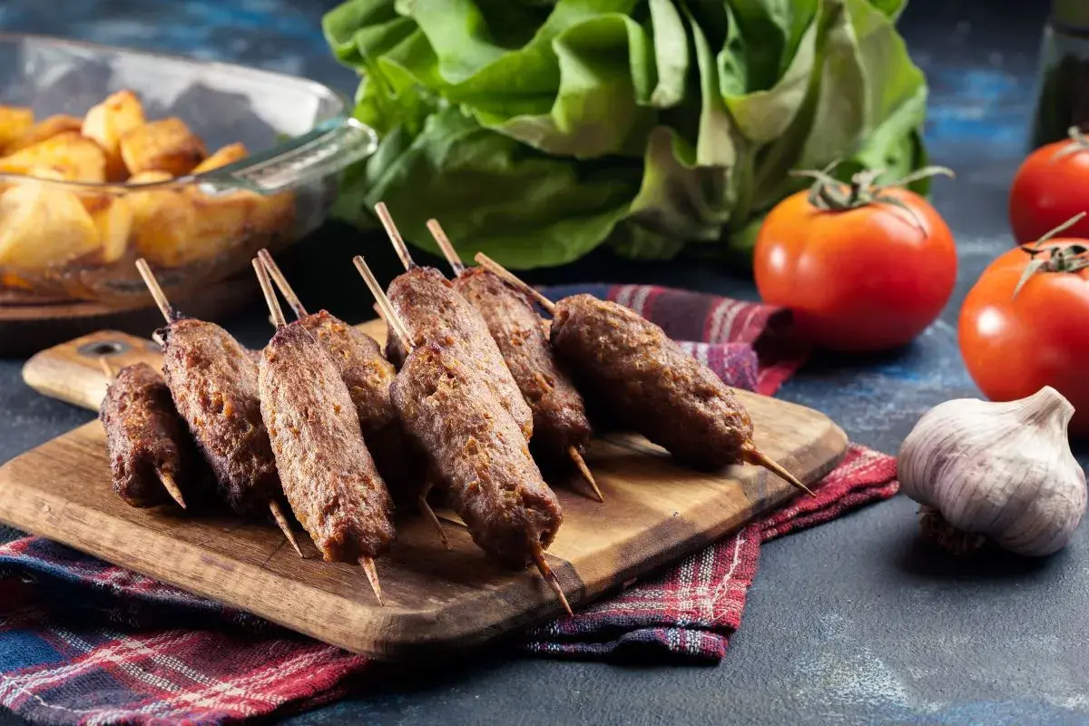 Grillowane ćevapi na drewnianej desce, obok pomidorów, czosnku i pieczonych ziemniaków.