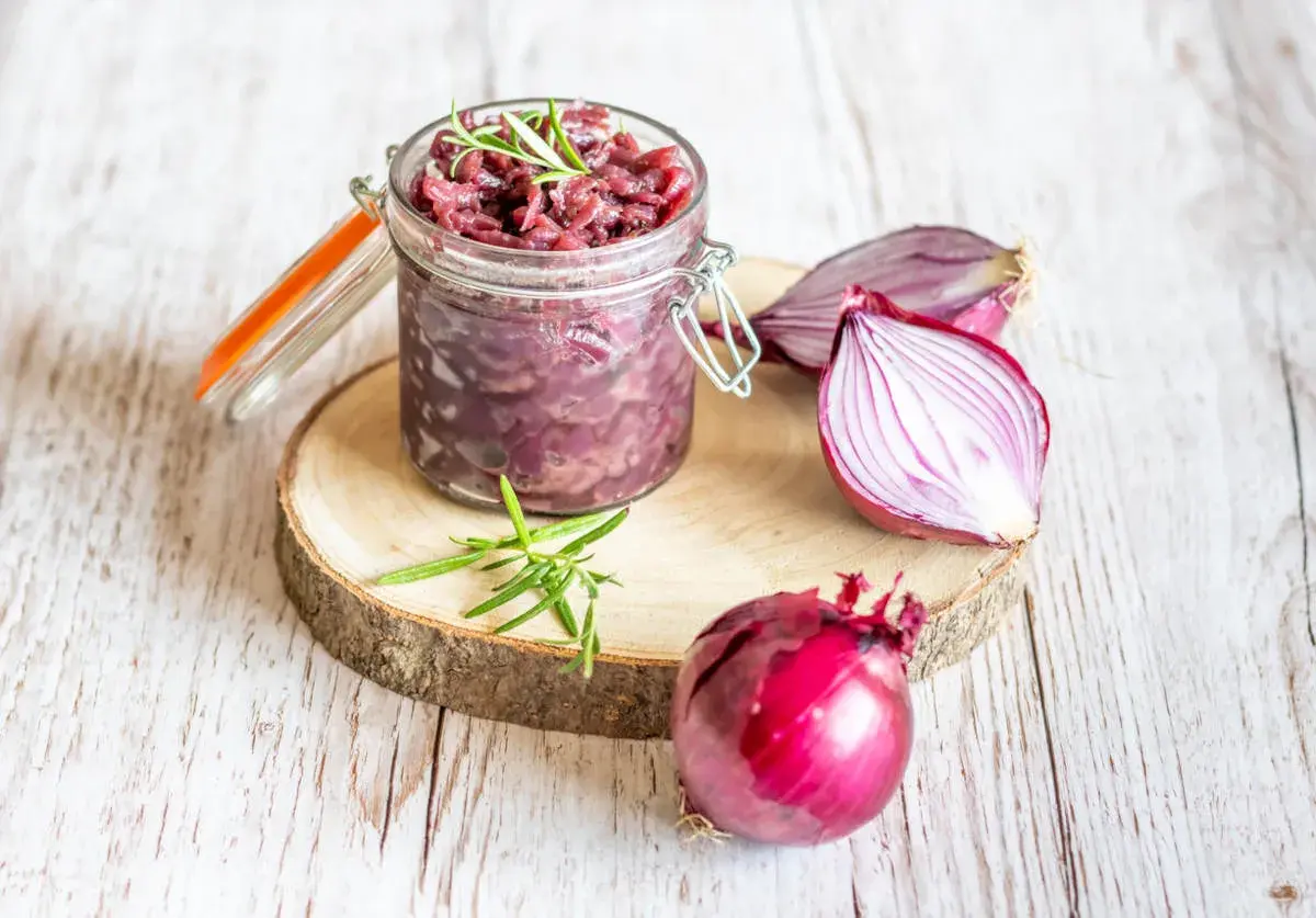 Słoik z konfiturą z czerwonej cebuli, obok cała cebula i połówka. Gałązka rozmarynu dodaje aromatu.