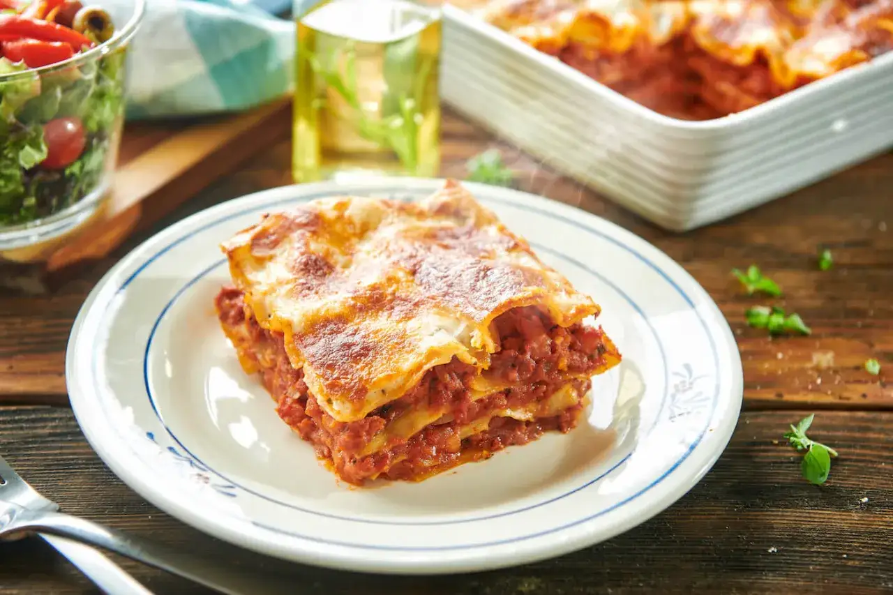 Pyszny kawałek lasagne, parujący z piekarnika, czeka na talerzu. Obok sałatka i oliwa.