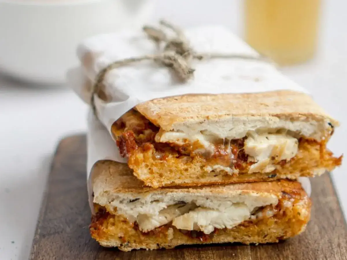 Dwa pyszne panini z kurczakiem, zawinięte w papier i przewiązane sznurkiem, leżą na drewnianej desce.