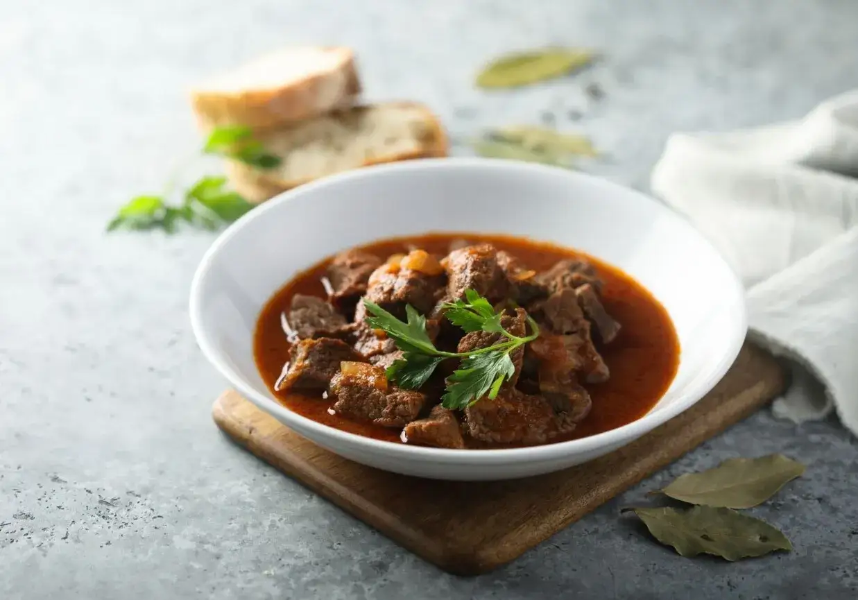 Aromaticzne mięso w sosie, podane w białej misce, ozdobione natką pietruszki. Obok kromki chleba i liście laurowe.