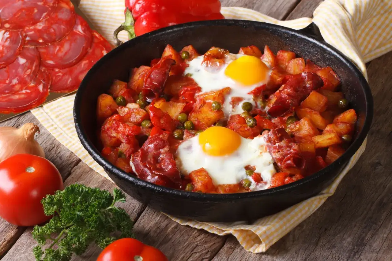 Pyszny obiad z jajek, ziemniaków i chorizo w patelni. Obok pomidory, cebula i natka pietruszki.