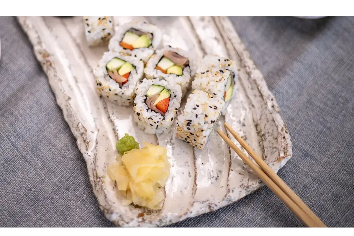 Uramaki z sezamem, awokado i ogórkiem na rustykalnym talerzu, obok wasabi i marynowanego imbiru.