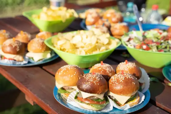 Mini burgery z serem i sałatą, miski chipsów i sałatki na piknikowym stole. Idealna szybka przekąska na letnie popołudnie.