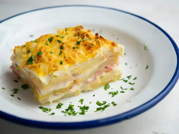 Kawałek pysznej, zapiekanej lasagne ziemniaczanej z szynką i serem, posypany natką pietruszki.