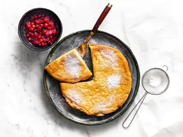 Puszysty omlet z air fryera, posypany cukrem pudrem, z łyżką żurawiny i sitkiem do cukru.