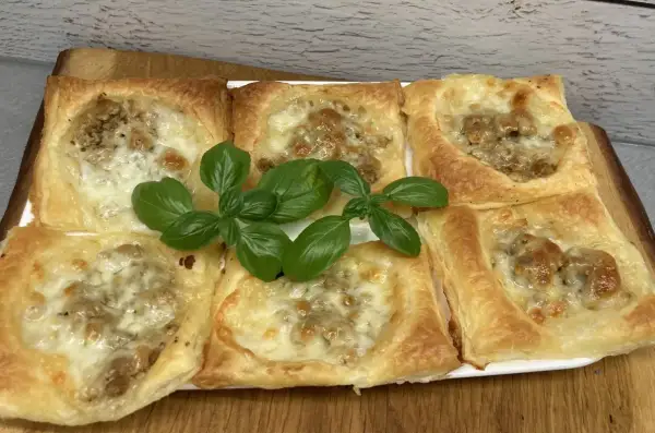 Sześć kwadratowych porcji ciasta francuskiego z mięsem mielonym i serem, ozdobione świeżą bazylią.