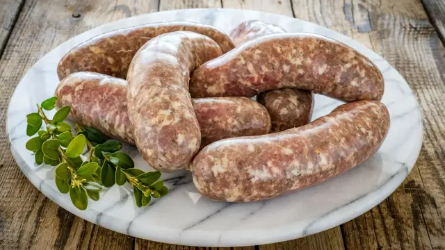 Surowa biała kiełbasa na marmurowej desce, z gałązką zieleni. Dowiedz się, ile czasu gotować białą kiełbasę, by była idealna.